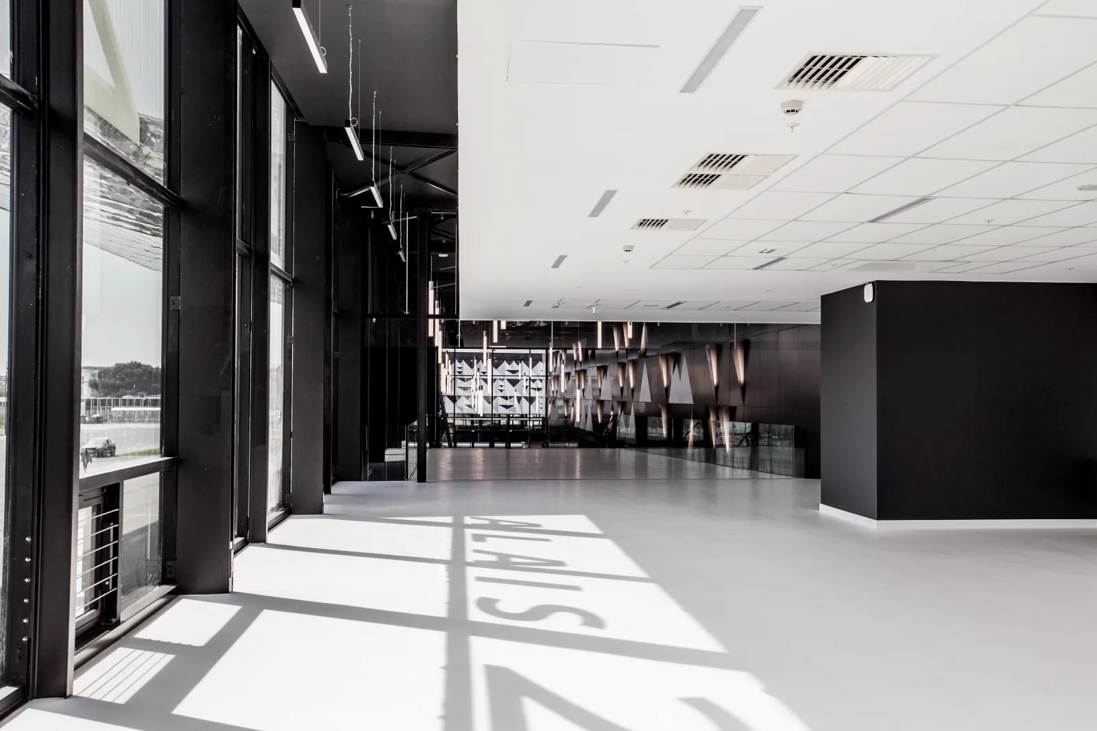 Modern interior of Palais 2 l'Atlantique in Bordeaux featuring a spacious empty hall with white polished floors, black structural elements, floor-to-ceiling windows, recessed ceiling lights, and natural daylight casting geometric shadows across the space.