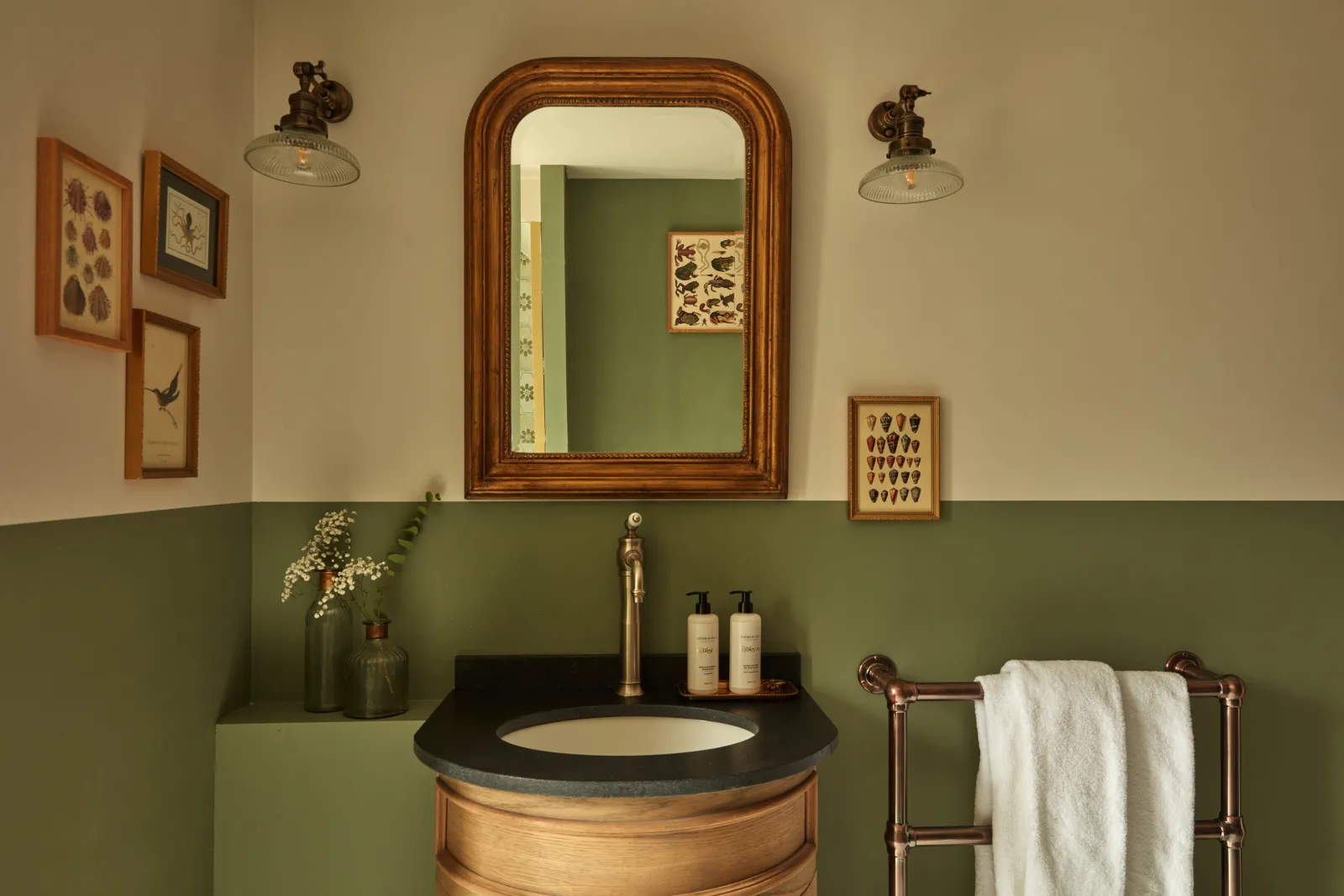 Elegant bathroom at Château Léognan featuring a round wooden sink with black countertop, ornate wooden mirror, vintage brass faucet, botanical framed artworks on sage green walls, and a heated towel rail with white towels.