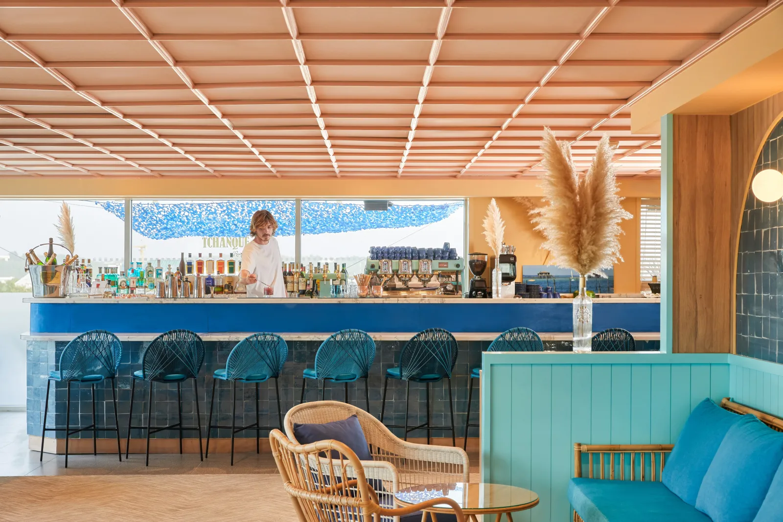 Modern bar lounge at Indigo Hotel Bordeaux with blue bar counter, turquoise bar stools, linear ceiling lights, pampas grass decoration, and comfortable seating area with wicker chairs