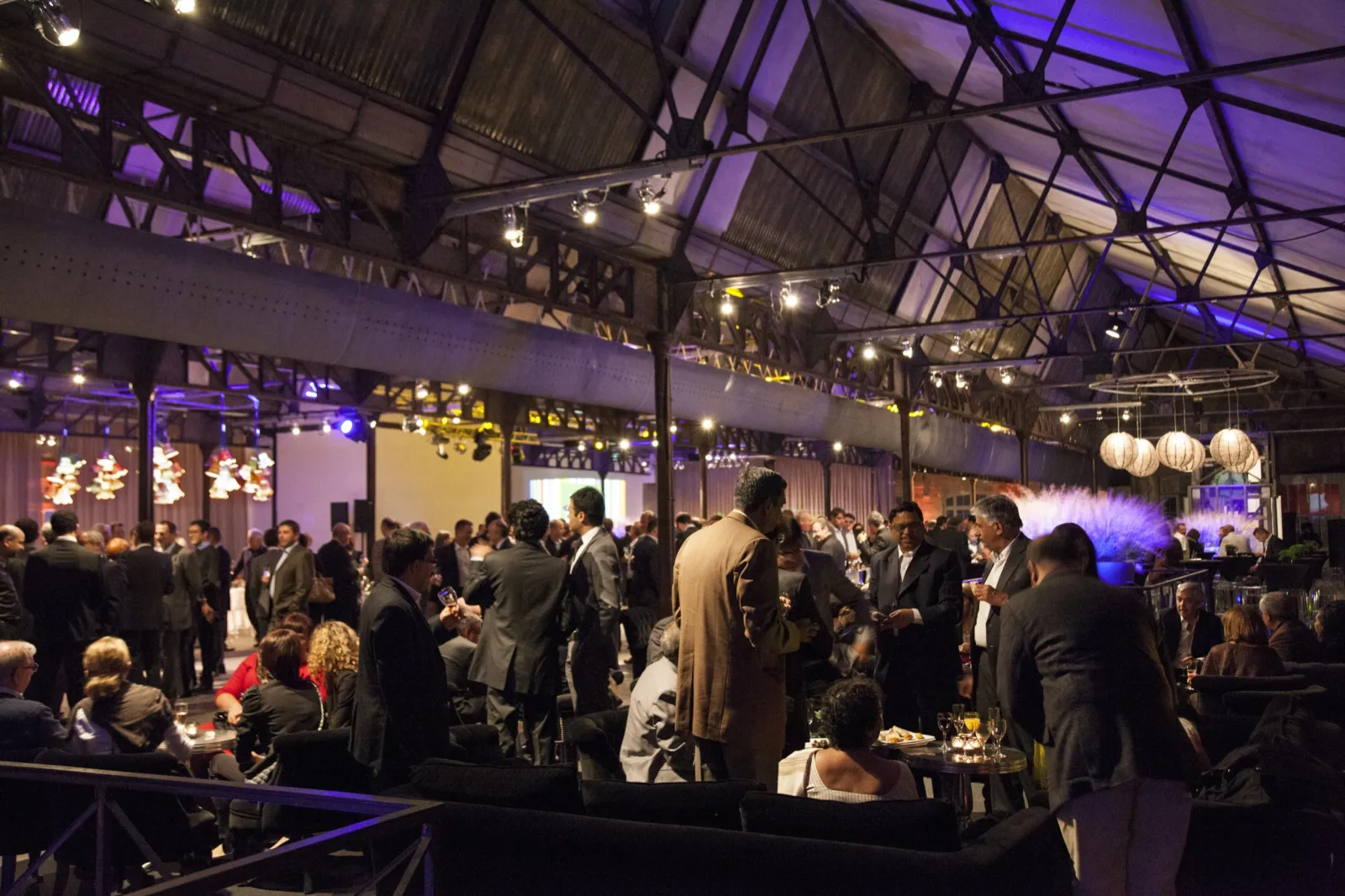 An elegant evening event at La Faïencerie in Bordeaux, featuring guests in formal attire mingling in a spacious industrial venue with purple ambient lighting, hanging lanterns, and exposed ceiling architecture.