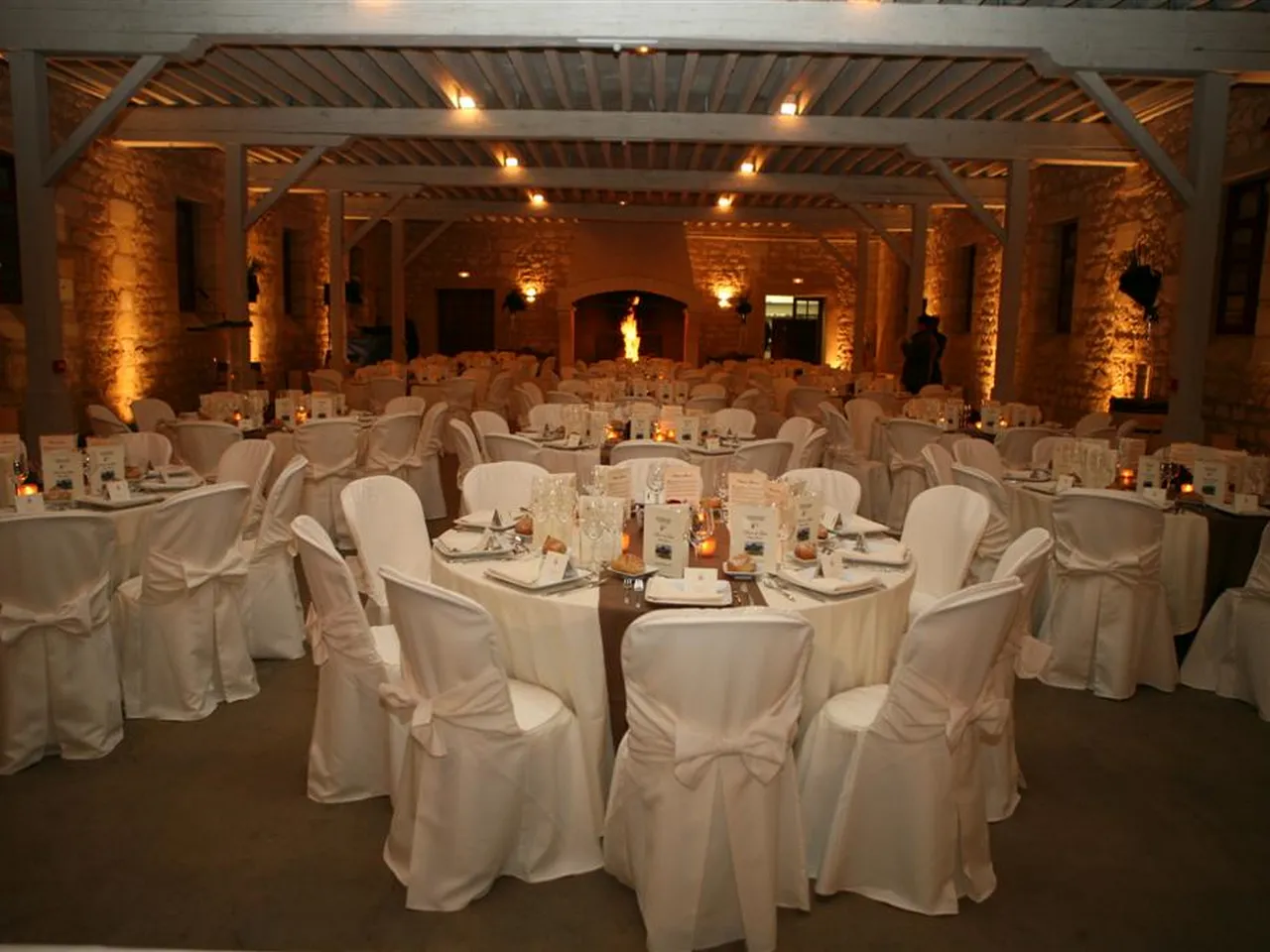 Elegant banquet hall at Château Giscours in Bordeaux with round tables dressed in white linens, white chair covers with bows, candlelit centerpieces, and warm amber uplighting on exposed brick walls with wooden beams.