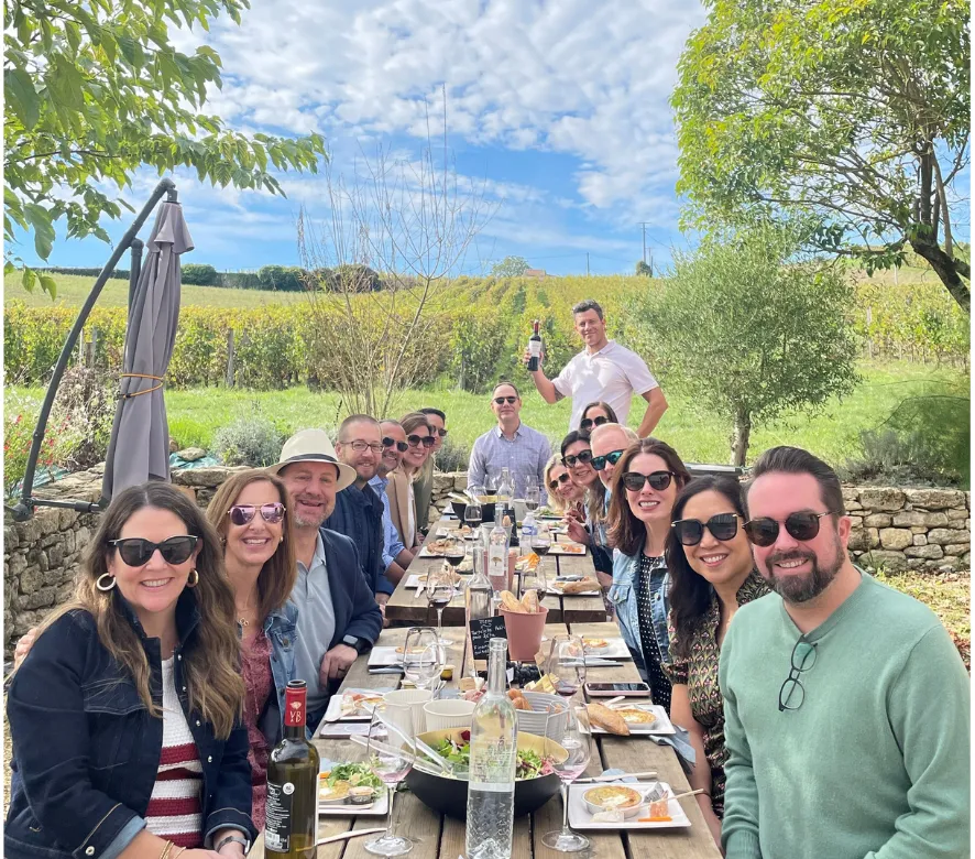 A group of people enjoying an outdoor lunch at a long wooden table in a Bordeaux vineyard, with rolling grapevines and countryside landscape in the background under a blue sky.