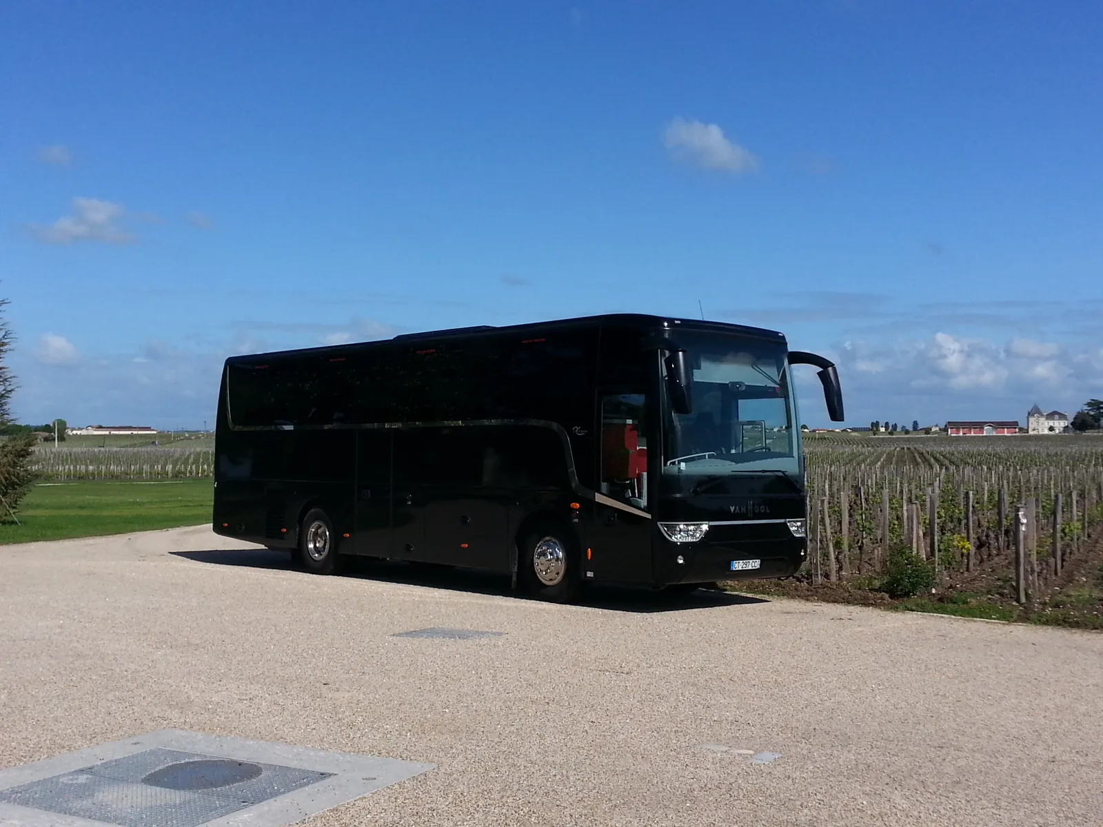 A black coach bus parked on a gravel road next to vineyards in the Bordeaux wine region, under a clear blue sky.