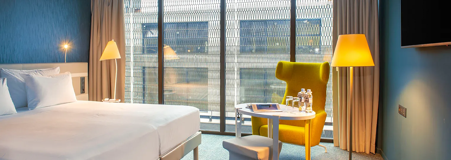 Modern hotel room at Radisson Blu Bordeaux featuring a bed with white linens, a yellow armchair with lamp, floor-to-ceiling windows overlooking the city, and contemporary furnishings in blue and neutral tones.