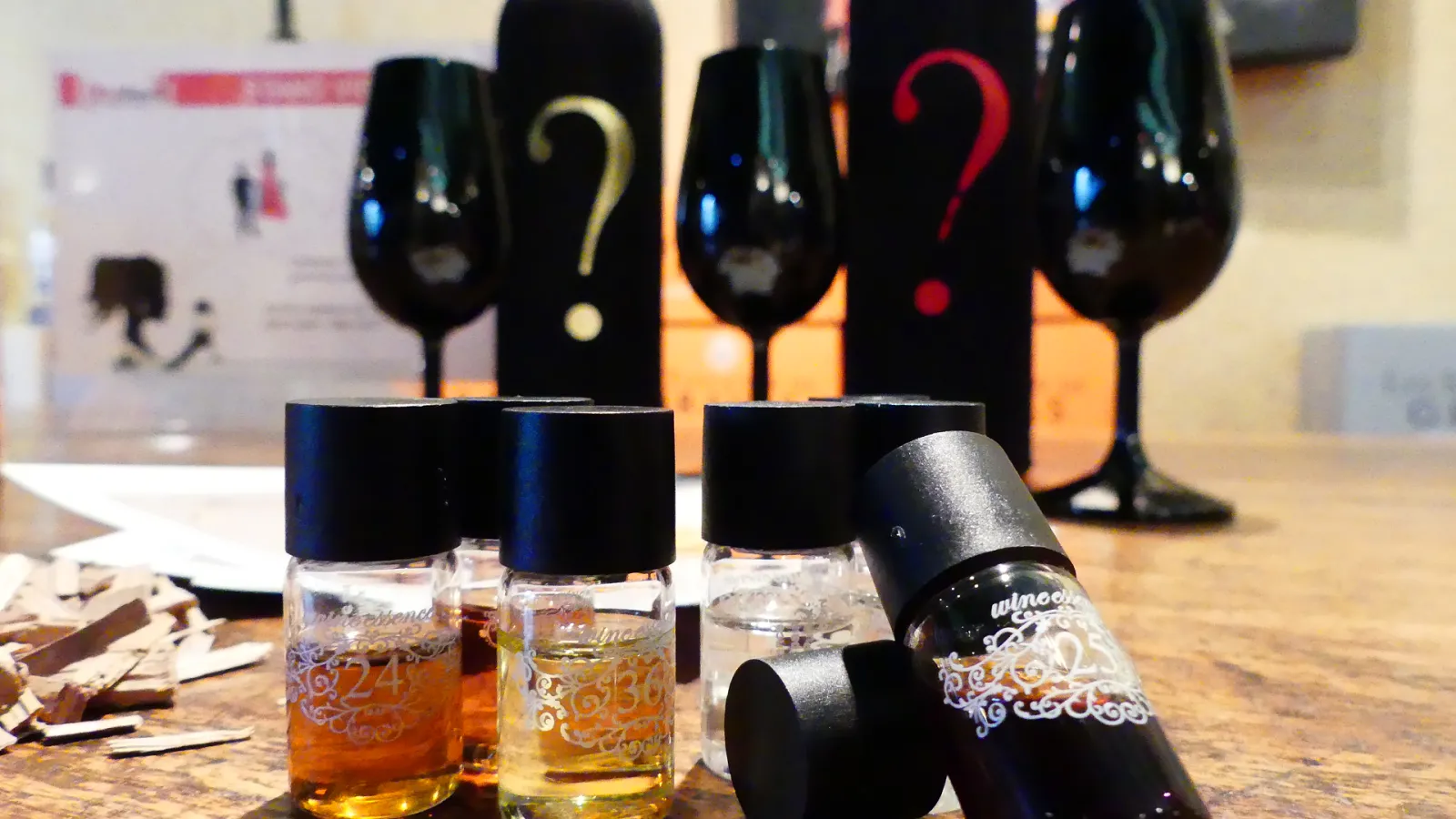 Small bottles of wine with black caps and decorative labels arranged on a wooden surface, with wine bottles displaying question marks in the background, suggesting a wine tasting or selection event in Bordeaux.