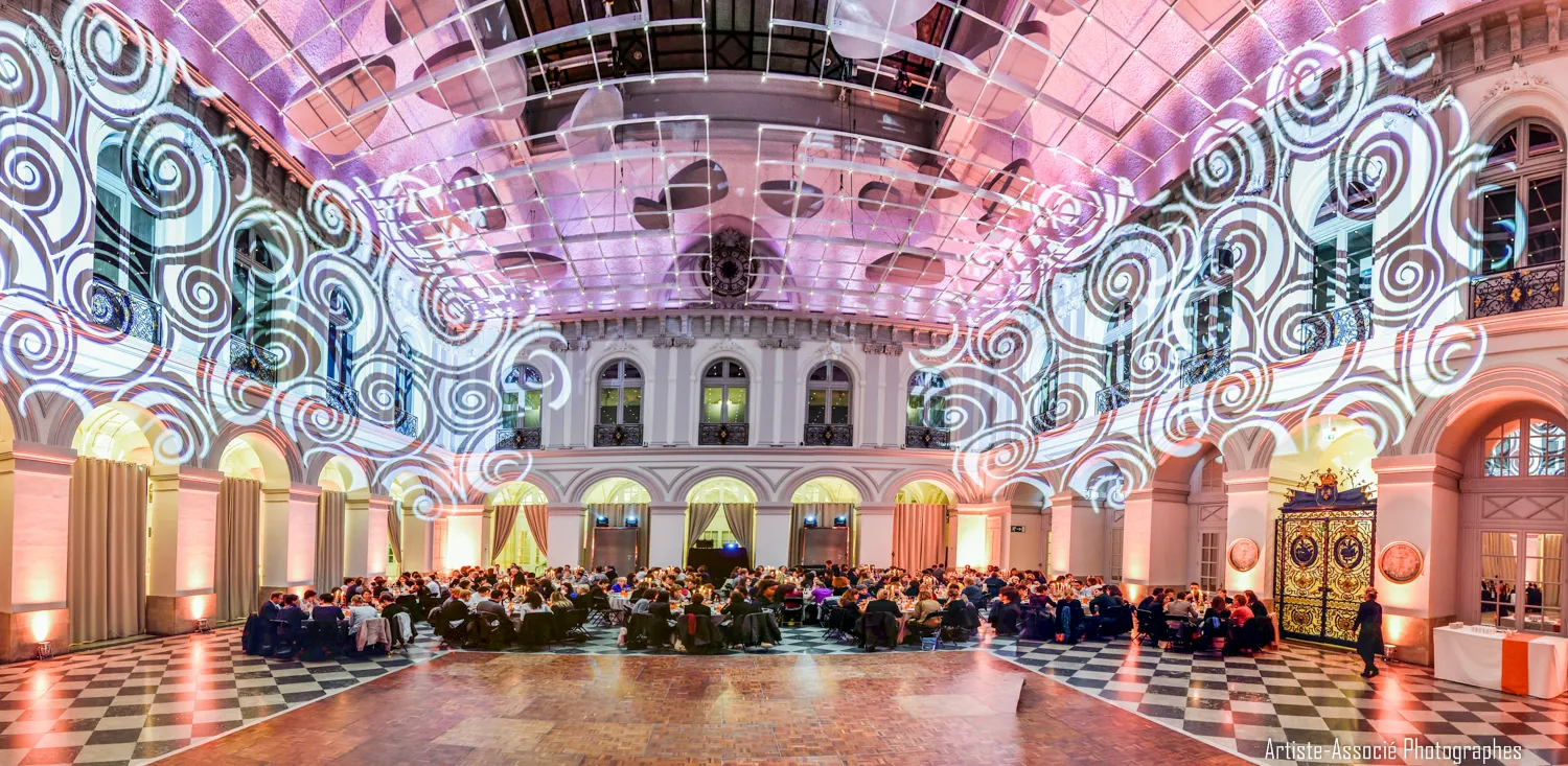 Grand ornate hall in Bordeaux's Palais de la Bourse with pink and purple lighting, spiral pattern projections on walls, classical architecture with arches, and a crowd of people gathered for an event.