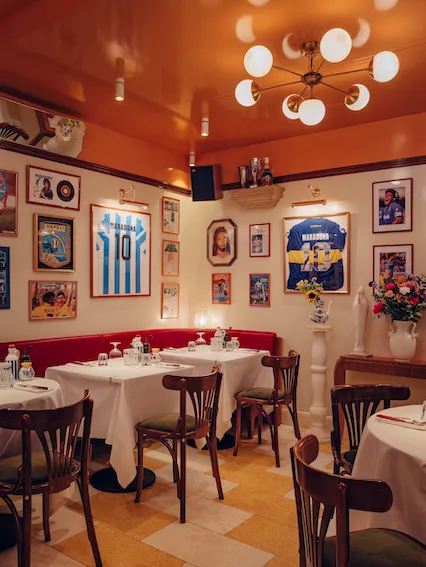 Elegant dining room at Osteria Palatino in Bordeaux featuring red banquette seating, white tablecloths, vintage football memorabilia on cream-colored walls, bentwood chairs, and a modern chandelier with warm orange ceiling lighting.