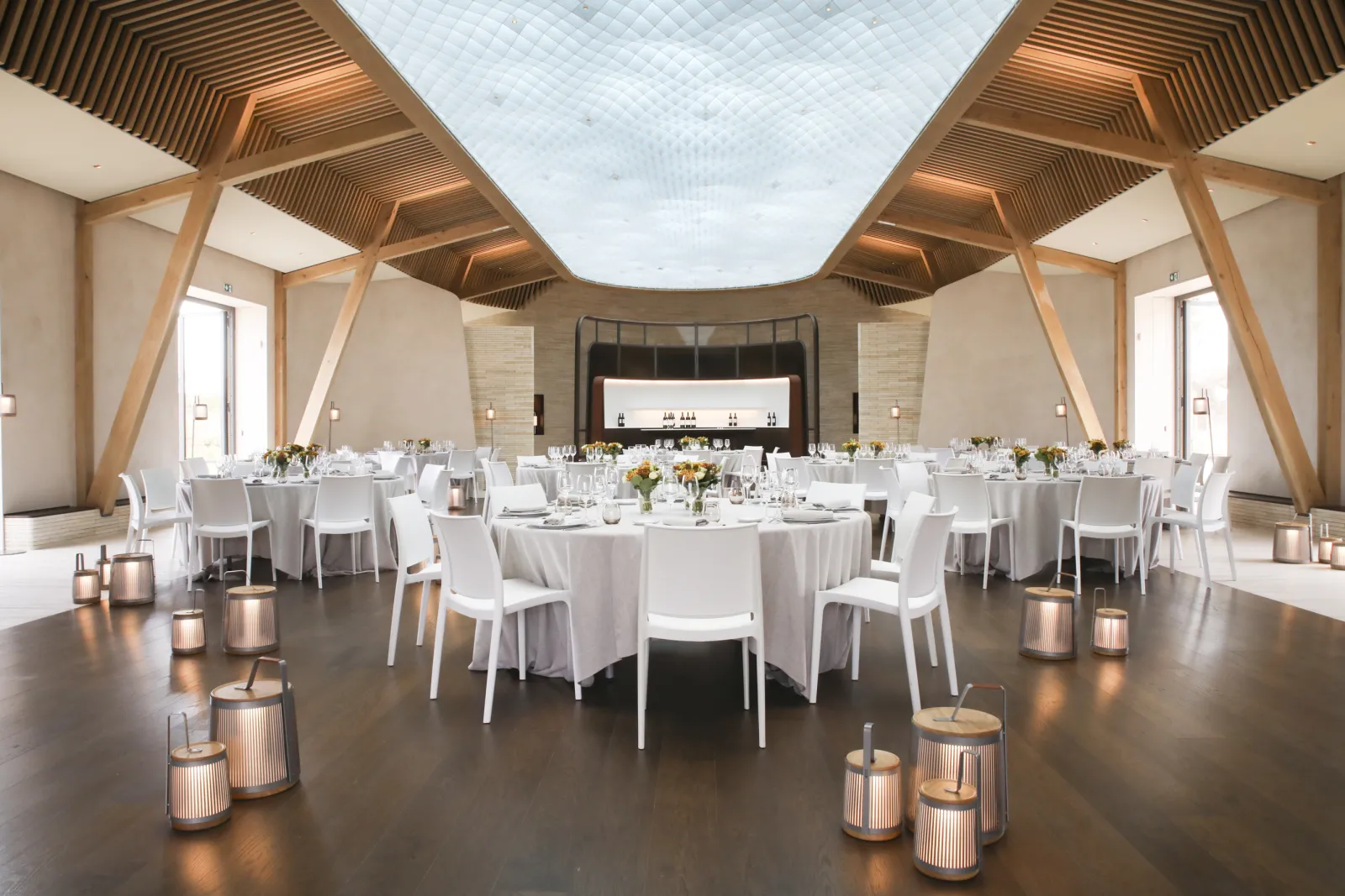 Elegant banquet hall at Château de Ferrand in Bordeaux with round tables set with white linens, modern wooden ceiling with skylights, and contemporary white chairs arranged for a formal event