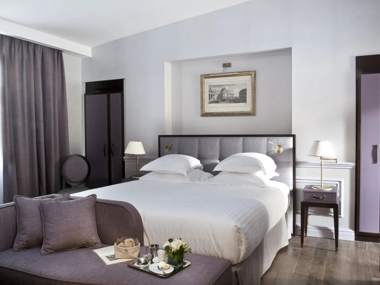 Modern luxury hotel bedroom at Hôtel de Sèze in Bordeaux, featuring a queen bed with gray upholstered headboard, white linens, twin nightstands with brass lamps, gray curtains, purple accent seating, and classical architectural framed print on the wall.
