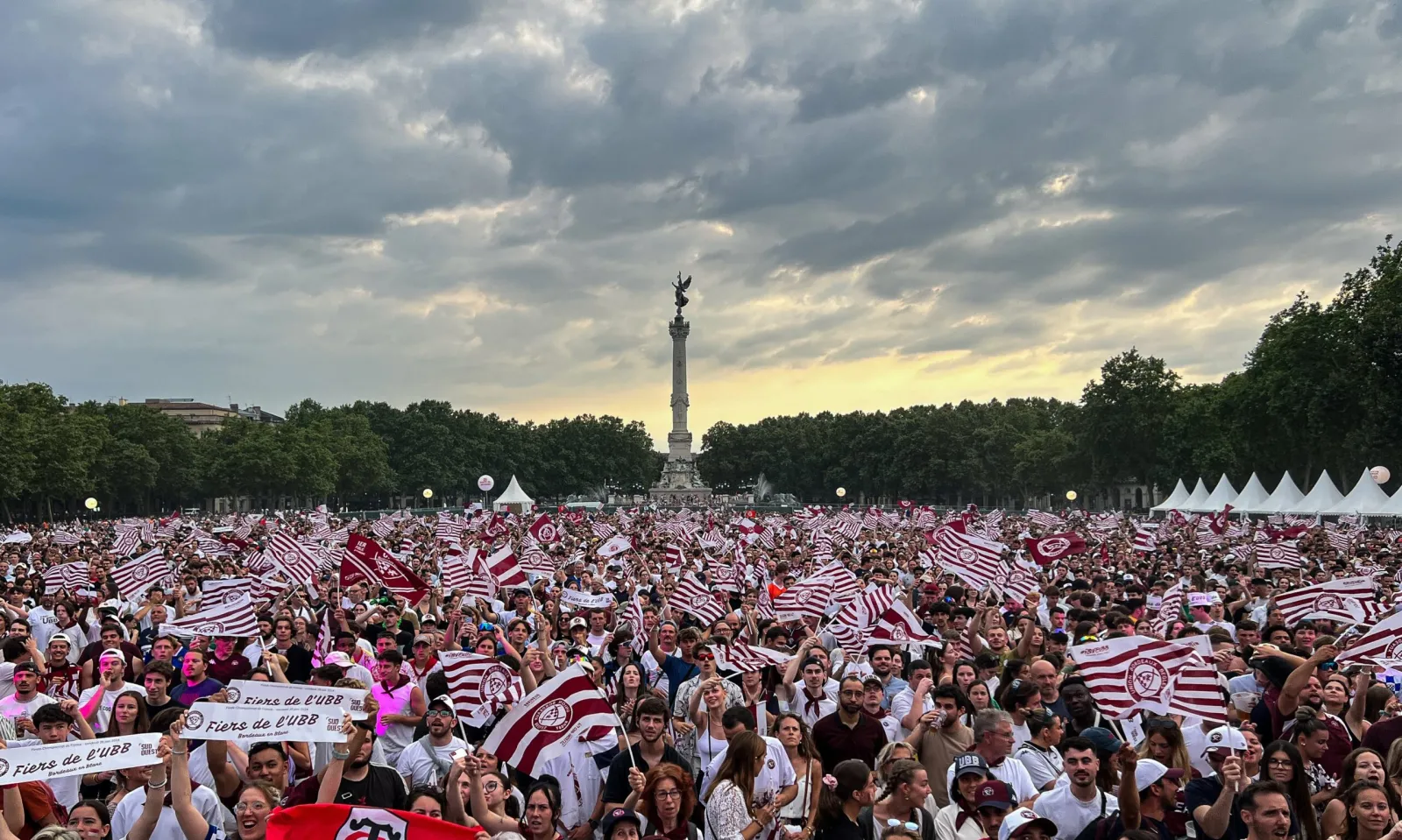 Large crowd of football supporters gathered at Esplanade des Quinconces in Bordeaux, waving red and white striped flags and banners, with a monument visible in the background under cloudy skies.
