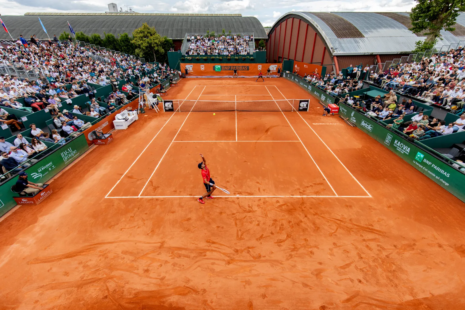 Professional tennis court with orange clay surface surrounded by spectators in stands, featuring BNP Paribas branding and traditional farm buildings in the background.