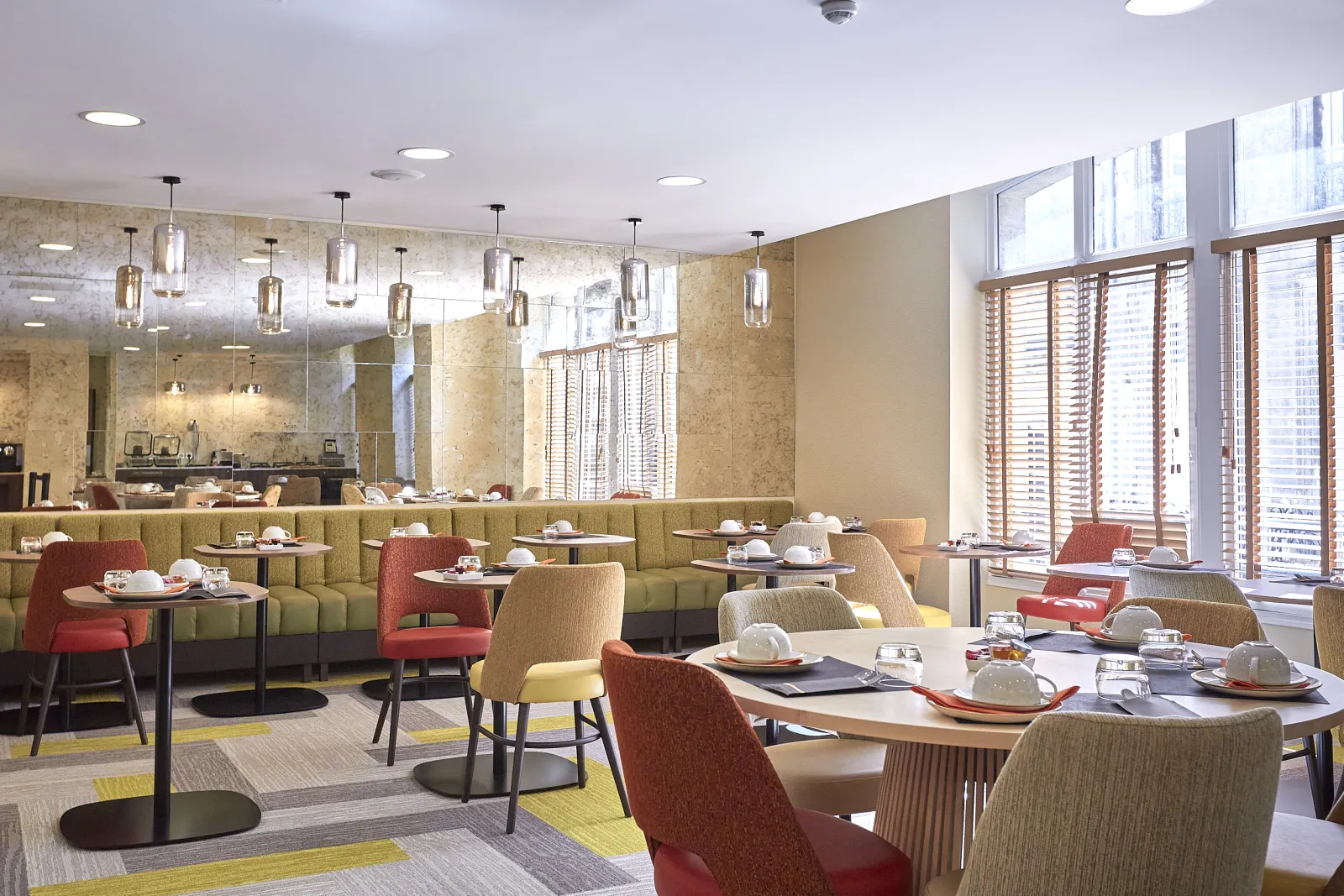 Modern hotel restaurant dining area with green banquette seating, red chairs, pendant lighting, floor-to-ceiling windows with wooden shutters overlooking the city, and tables set with white tableware.