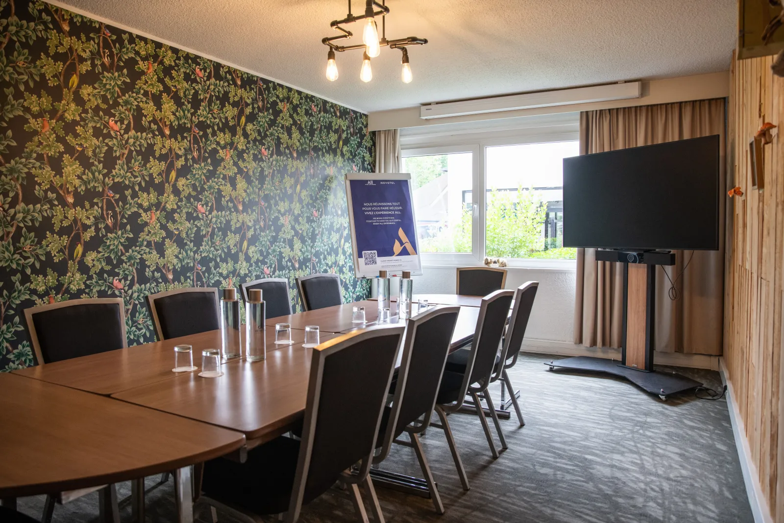 Modern conference room at Novotel Bordeaux Mérignac with wooden table, black chairs, floral patterned wallpaper, large display screen, and natural light from windows