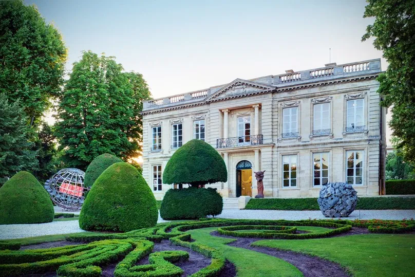 Elegant neoclassical mansion in Bordeaux with manicured topiary gardens, featuring sculpted hedges, a decorative metal sphere sculpture, and ivy-covered walls framing the formal residence.