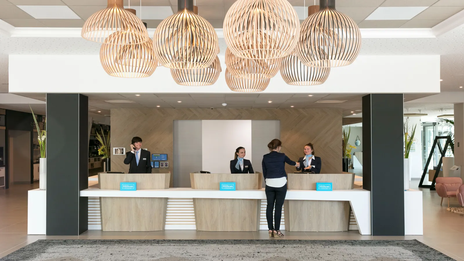 Modern hotel reception desk of Hilton Garden Inn Bordeaux Centre with staff members assisting guests, featuring wooden counters, pendant lamps, and contemporary interior design.