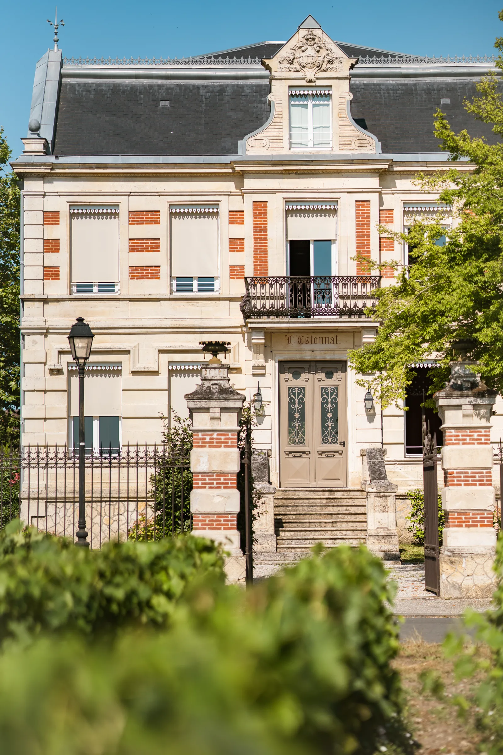 Front facade of a 19th-century Haussmann-style mansion in Bordeaux featuring cream and red brick walls, mansard roof with dormer window, ornate entrance gate, and wrought iron balcony, surrounded by green foliage.