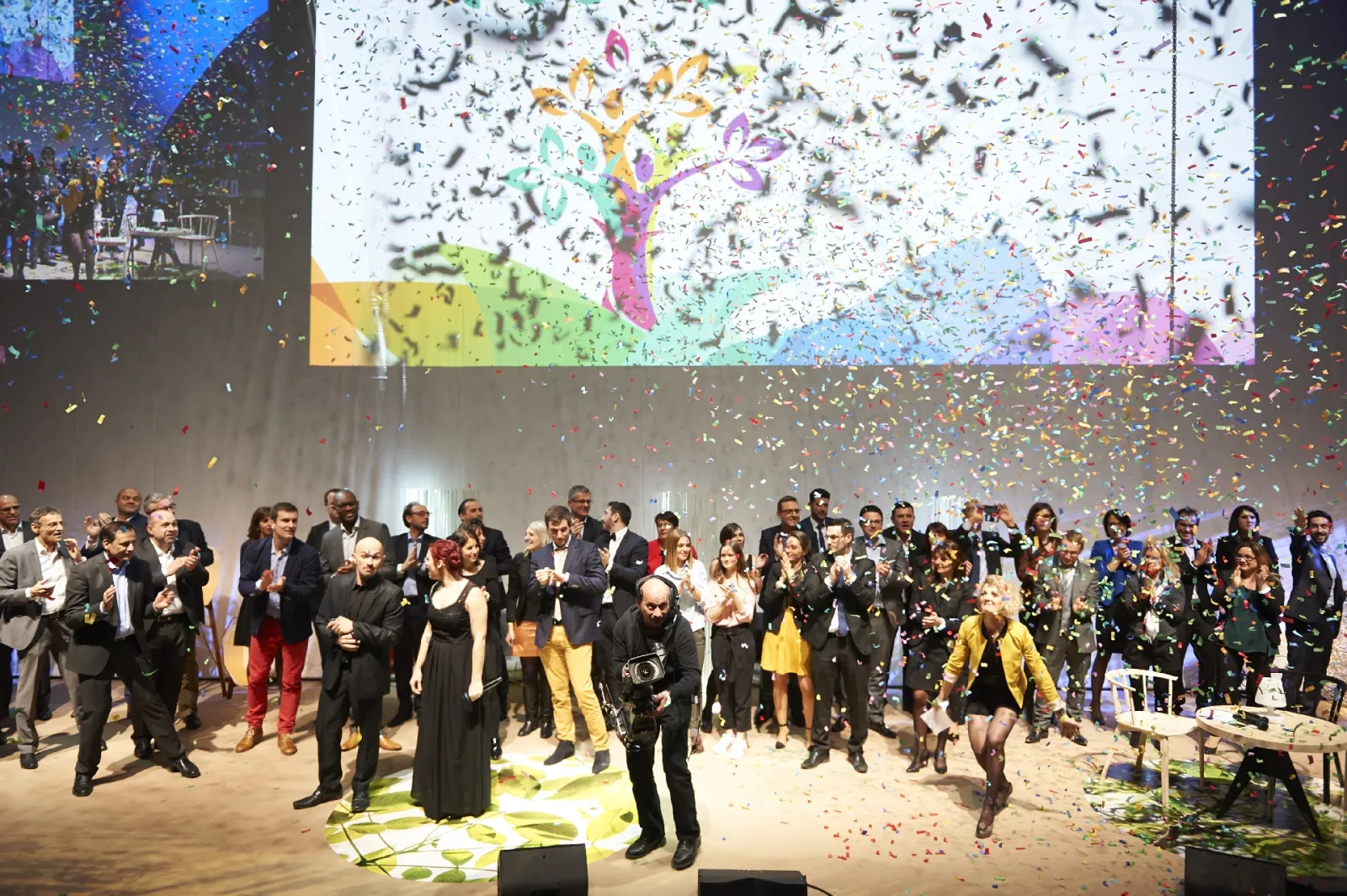 Large indoor celebration event with confetti falling on a diverse crowd of formally dressed attendees in front of a colorful projection screen displaying a stylized tree design