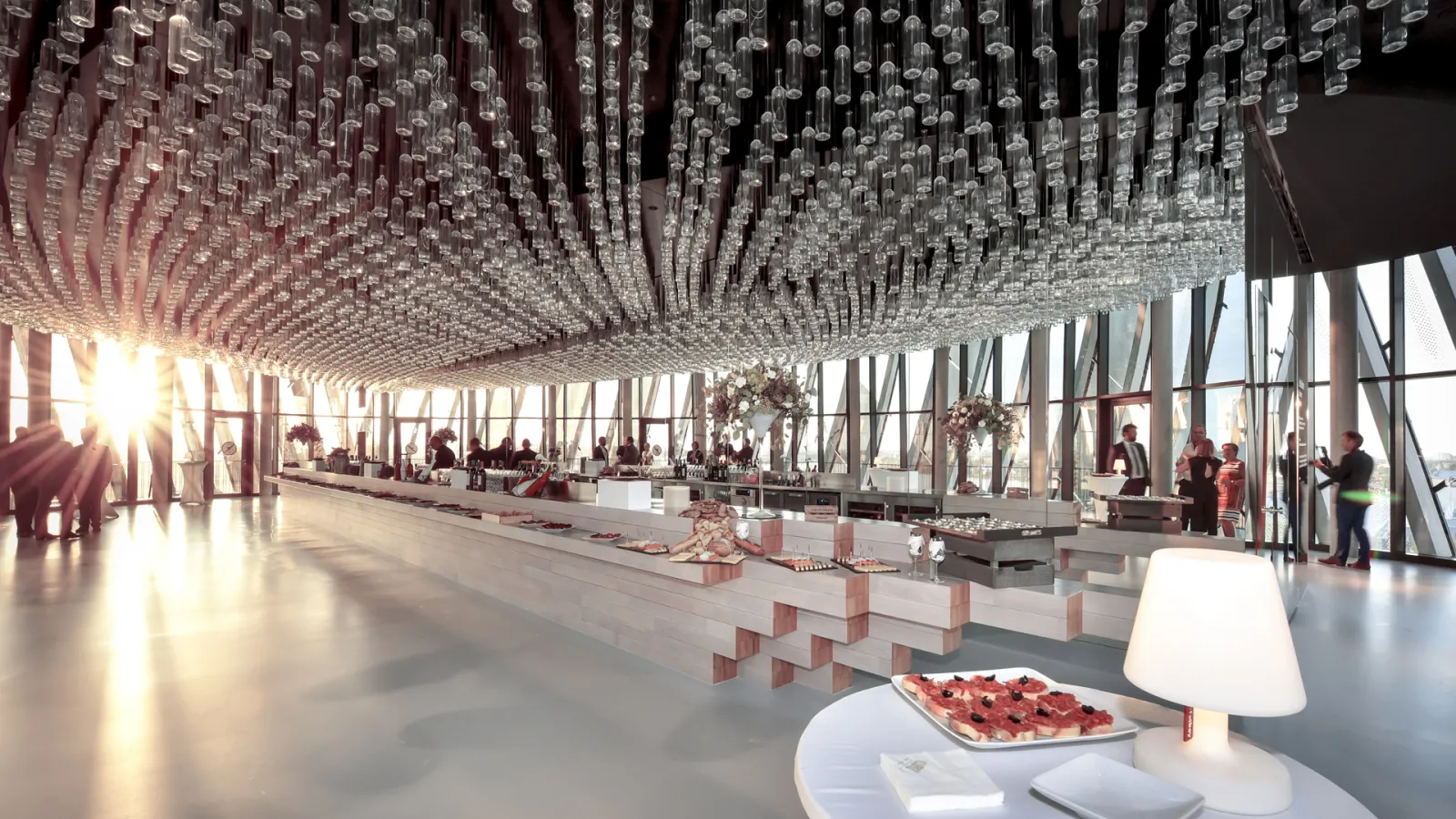 Modern interior of Cité du Vin in Bordeaux featuring a spacious venue with thousands of hanging glass bottles as ceiling decoration, floor-to-ceiling windows, white furniture, and visitors exploring the exhibition space flooded with natural light