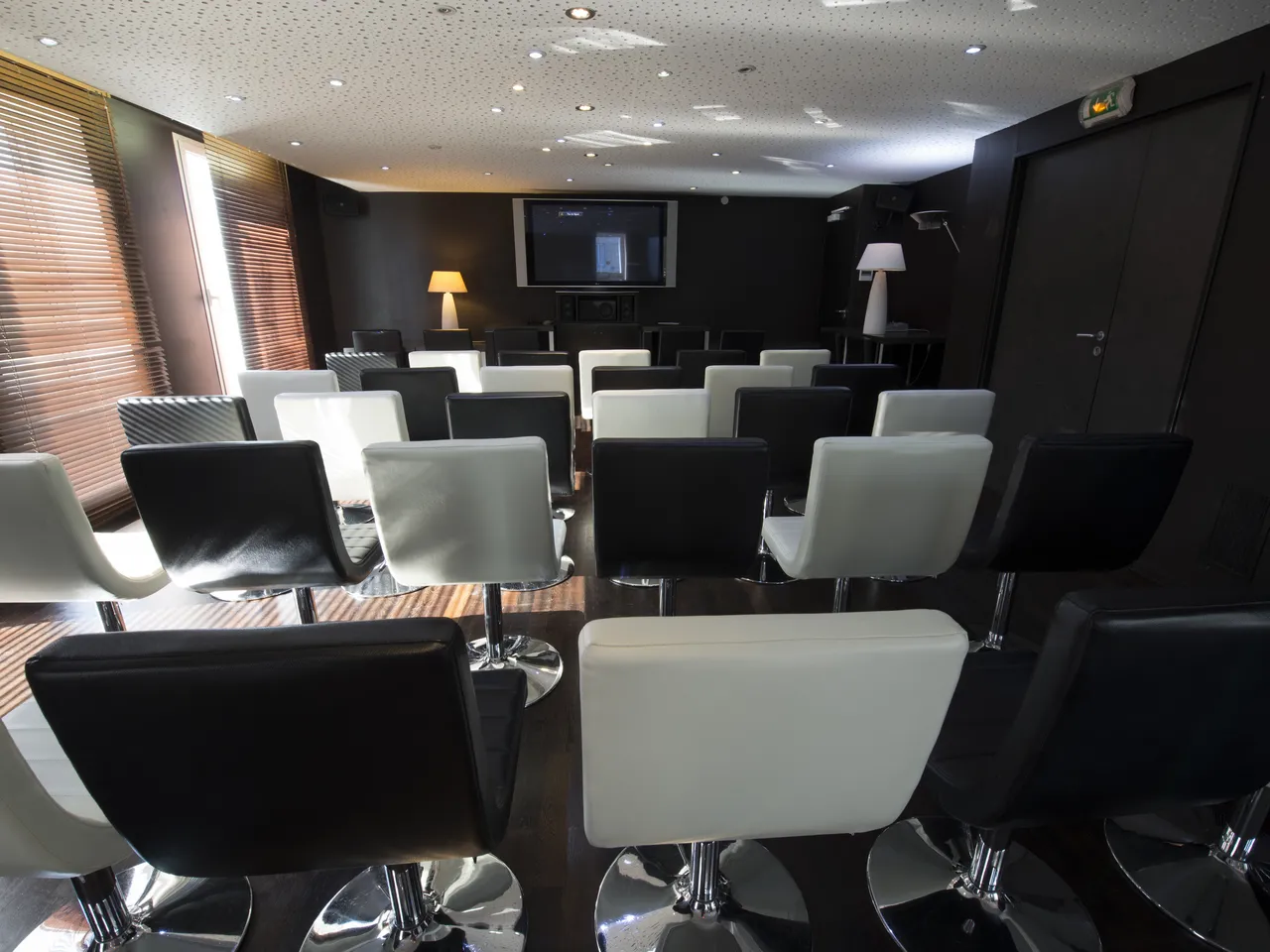 Modern cinema room at Seeko'o Hotel Bordeaux with alternating black and white comfortable chairs arranged in rows, ambient lighting, and a wall-mounted screen.