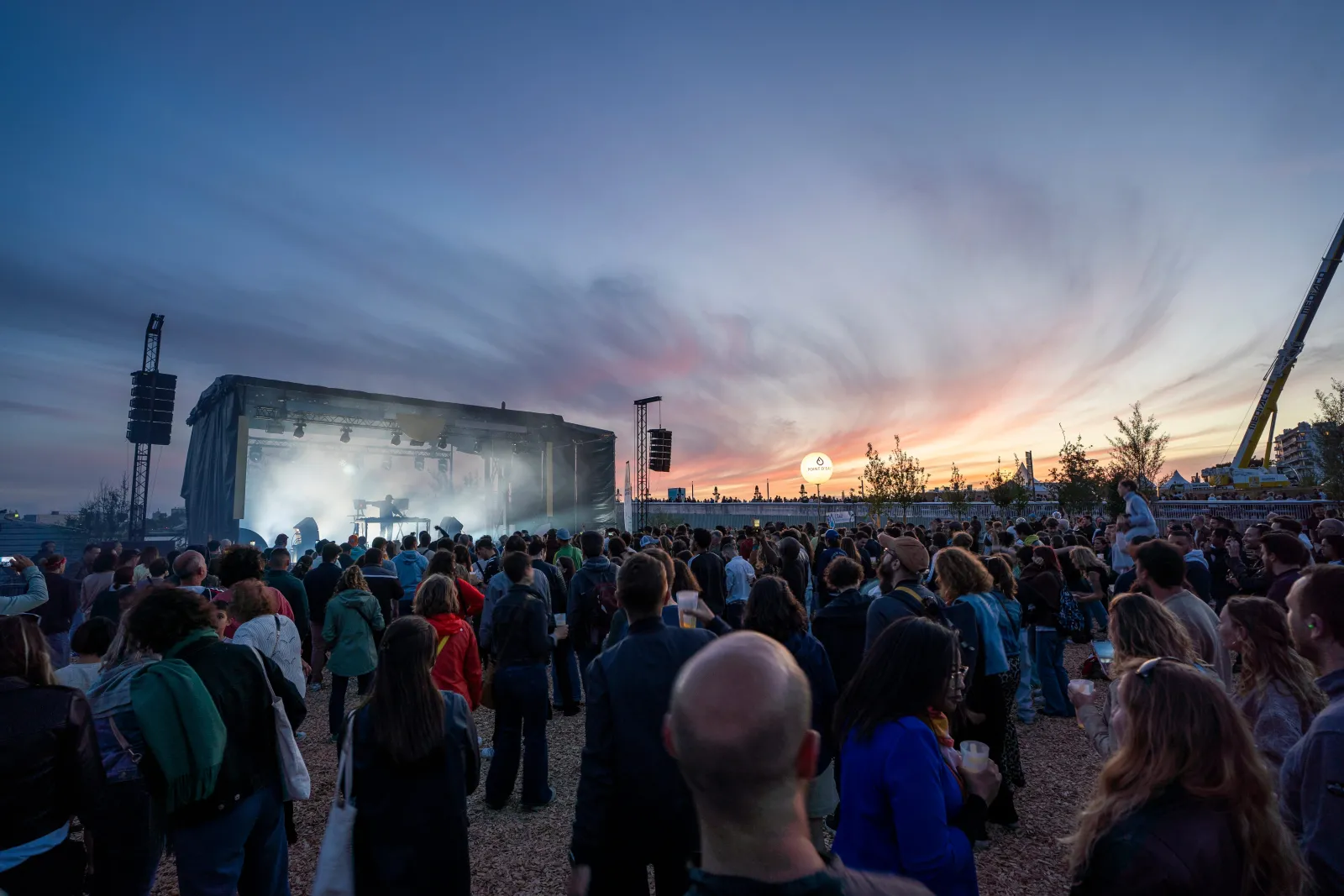 Large outdoor concert crowd gathered at dusk with illuminated stage and sound equipment, riverside setting in Bordeaux