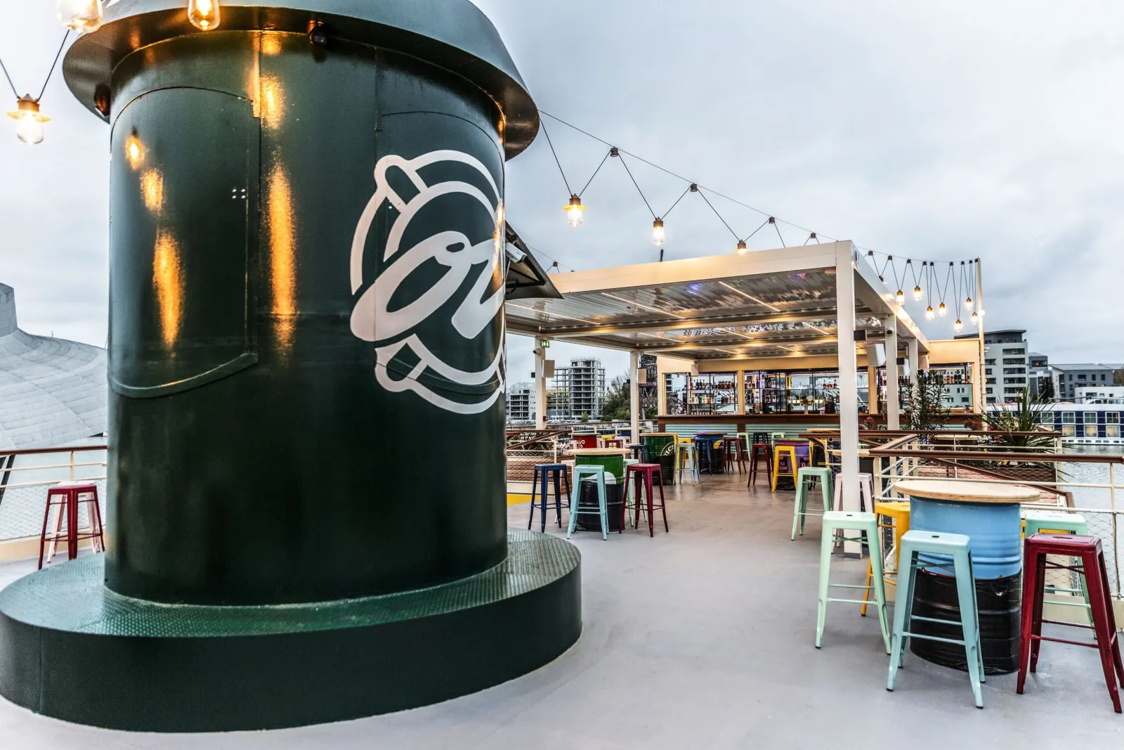 Outdoor rooftop bar and restaurant with a large green cylindrical structure featuring a white logo, colorful bar stools, wooden pergola with hanging lights, and waterfront views in Bordeaux