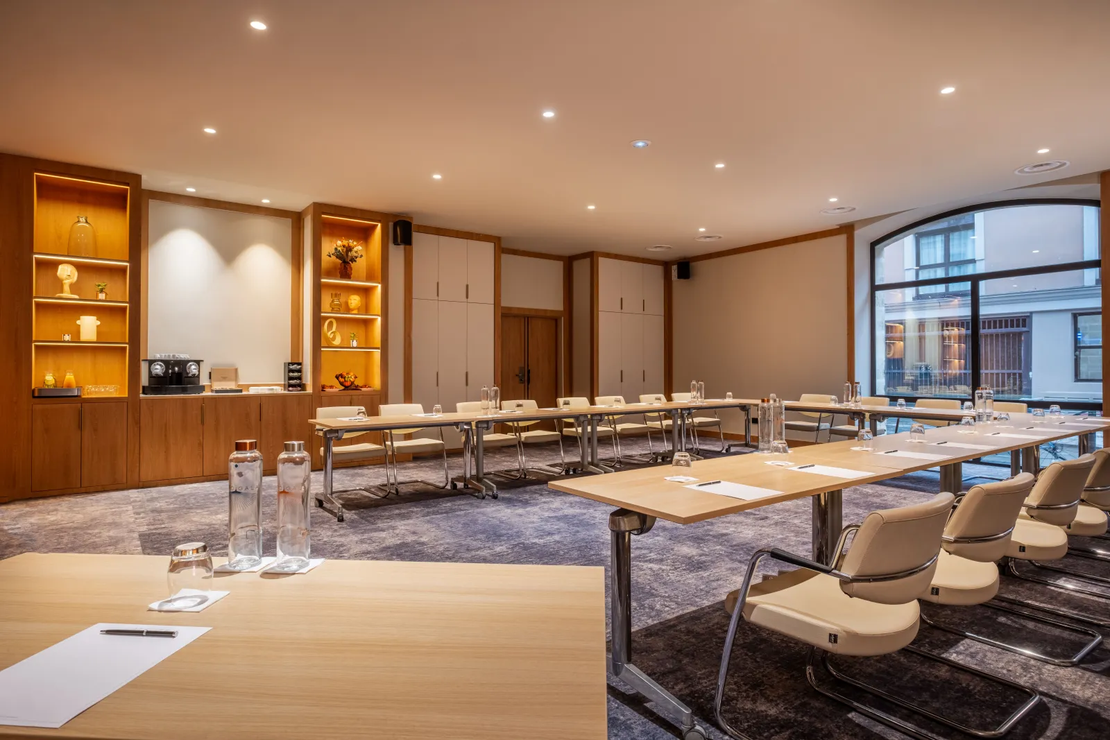 Modern conference room with multiple wooden tables, beige chairs, built-in shelving with warm lighting, and floor-to-ceiling windows overlooking Bordeaux streets.