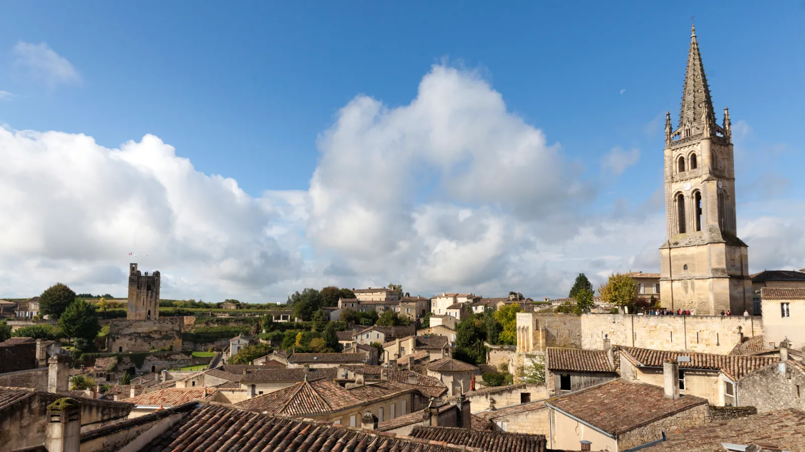 Panoramic view of a medieval French village with stone buildings, terracotta roofs, and a prominent church bell tower under a blue sky with white clouds.