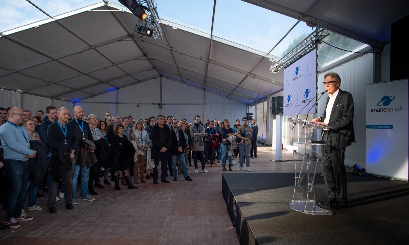 A man in a dark suit and glasses speaks at a podium under a white tent to a large audience at an outdoor event in Bordeaux, with Ariane Group branding visible.