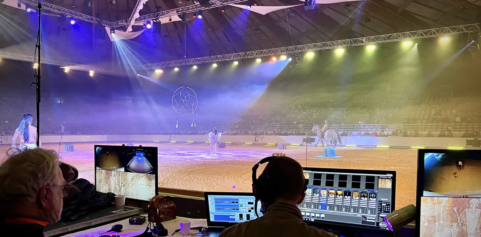 Control room operators manage a live circus performance under dramatic blue and green lighting, with acrobats performing in a sand arena beneath a large dreamcatcher prop and industrial stage rigging.