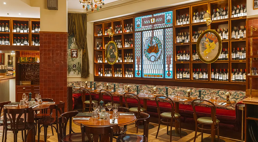 Elegant wine bar interior with wooden shelving displaying hundreds of wine bottles, red velvet banquette seating, round tables, ornate decorative panels, and vintage wine-themed artwork on the walls.