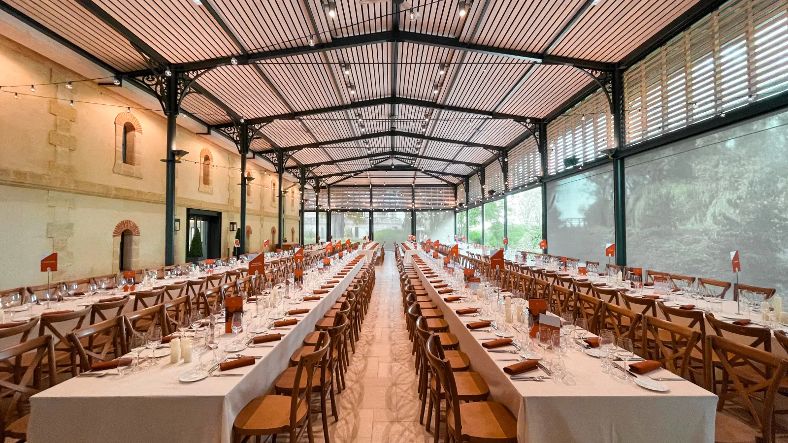 Large banquet hall with long white-draped tables, wooden cross-back chairs, and place settings under a modern steel and polycarbonate arched roof, stone walls visible on the left side