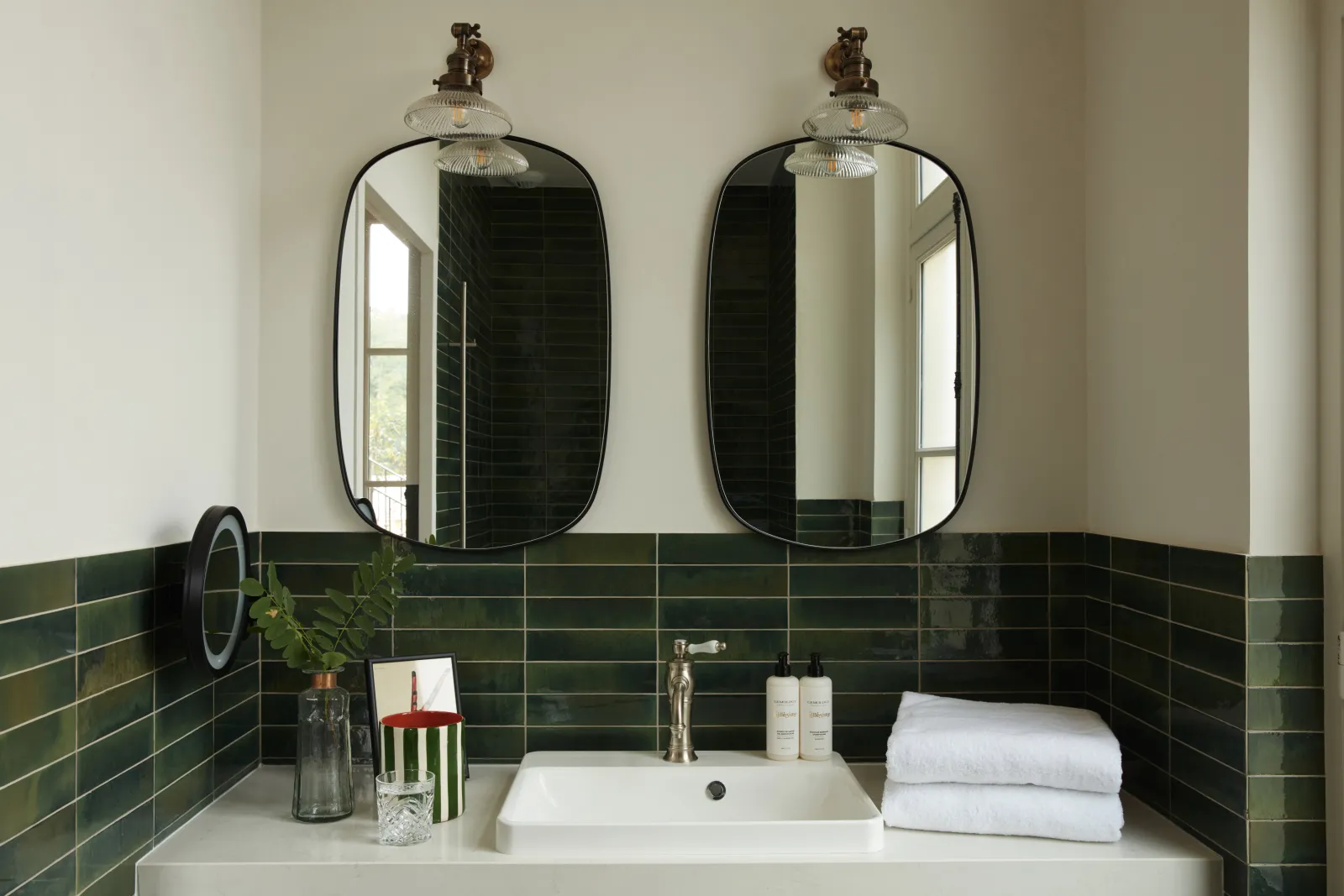 Modern bathroom vanity with dual rounded black mirrors, brass sconces, white sink, green subway tile backsplash, and toiletries at Château Léognan in Bordeaux