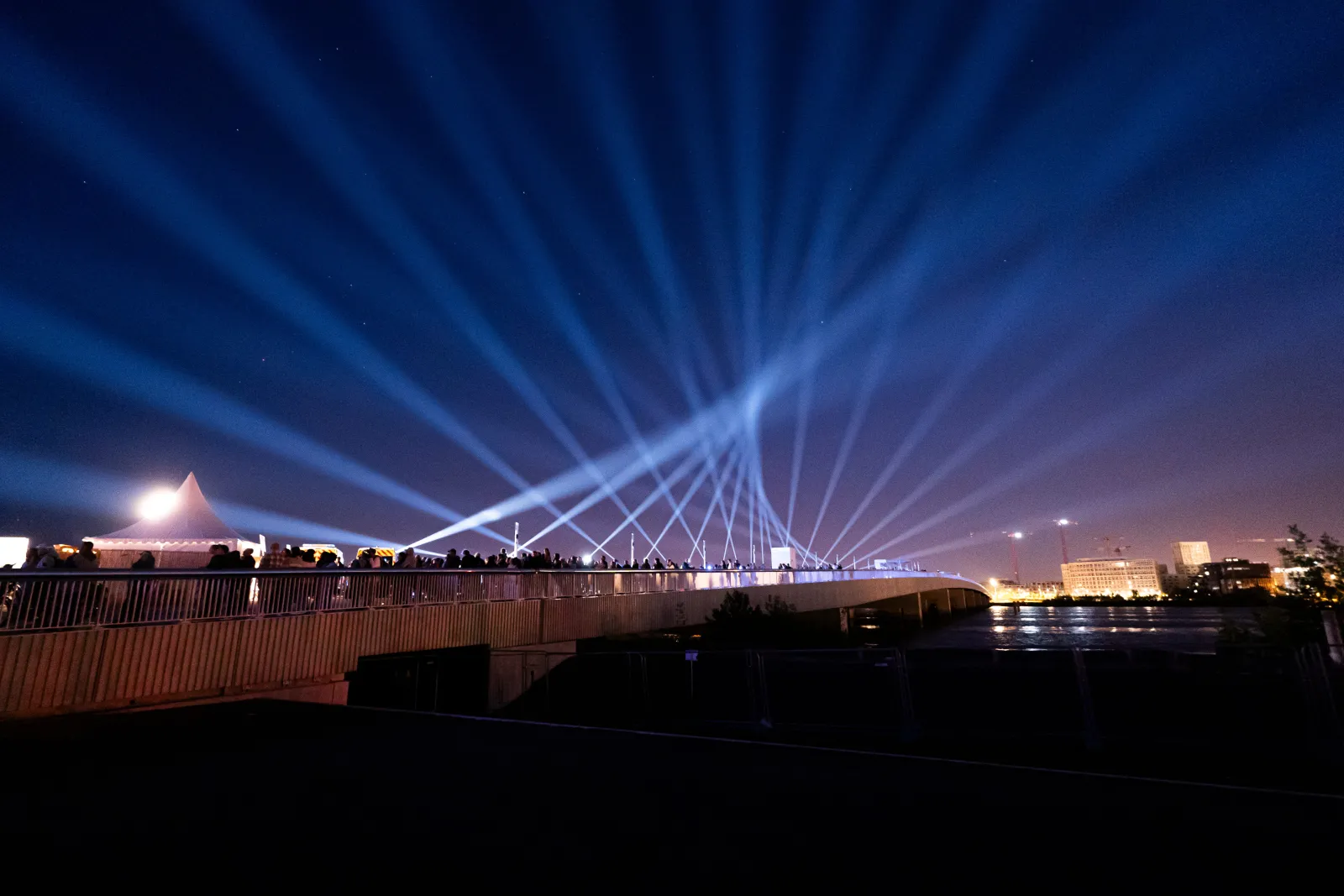 Illuminated modern bridge with dramatic light beams crossing overhead at night, spanning a river with city lights reflected in the water and buildings visible in the background.