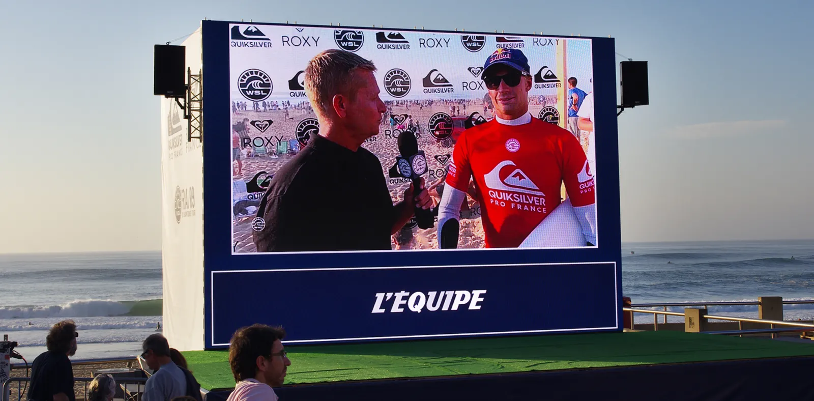 Large outdoor screen displaying a live interview at a professional surfing event, with a surfer in red Quiksilver gear being interviewed on a beach, with ocean and spectators visible in the background