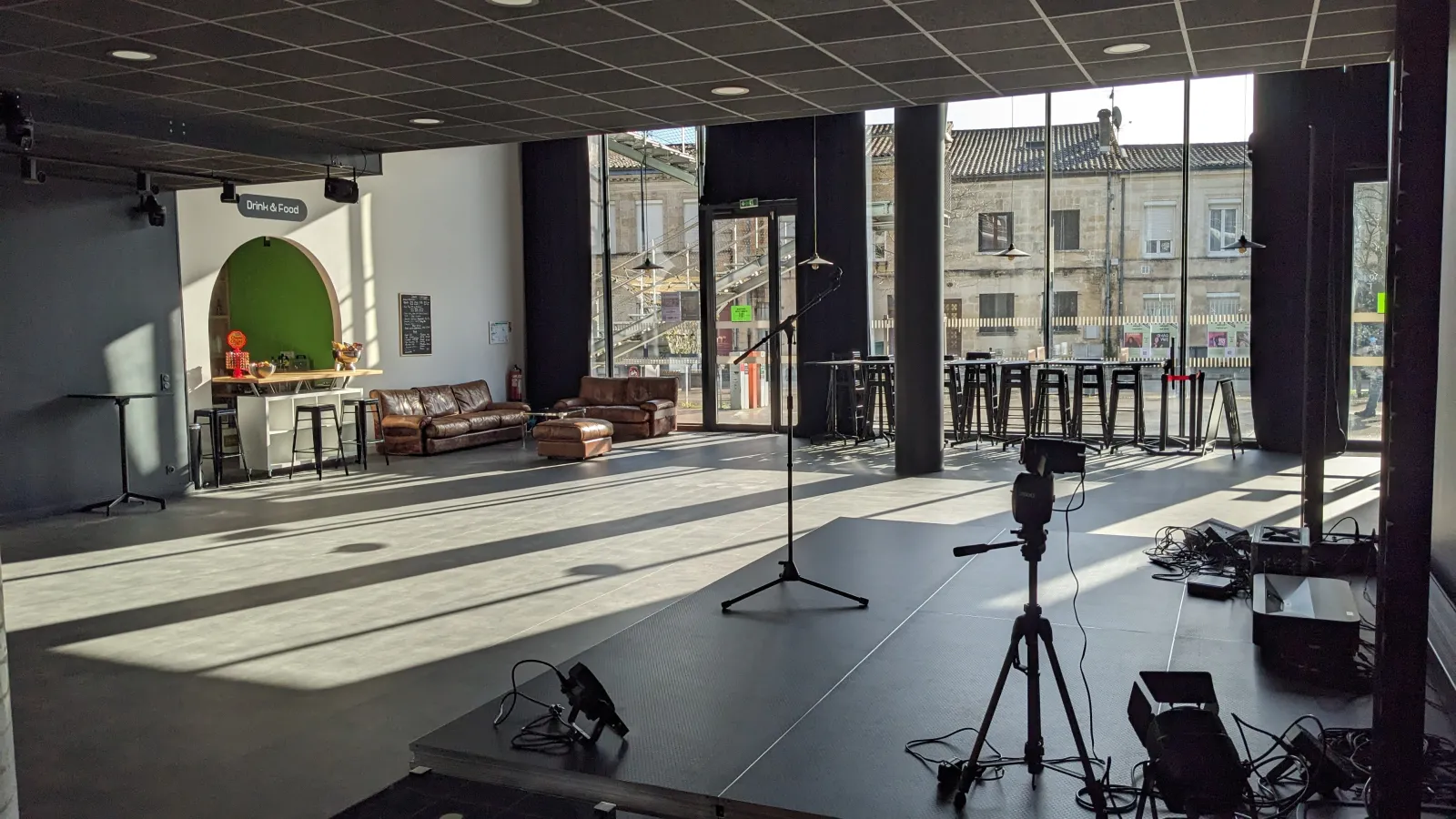 Modern event space at L'Estrade in Bordeaux with professional lighting equipment, brown leather seating area, green accent wall, bar counter, and large windows overlooking historic buildings