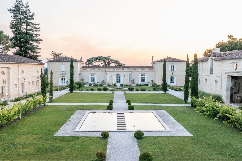 Elegant neoclassical estate with white stone buildings surrounding a manicured courtyard featuring a central rectangular pool, cypress trees, and manicured lawns at dusk in Bordeaux.