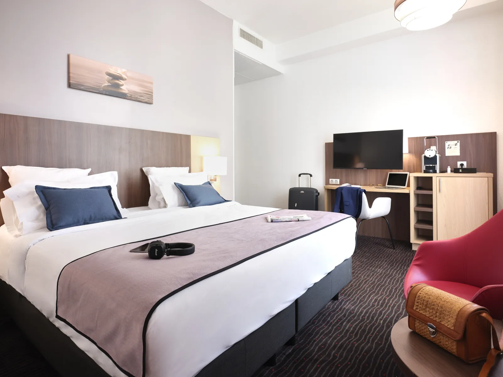 Modern hotel room at Quality Hôtel Bordeaux Centre featuring a queen bed with white linens and blue pillows, wooden headboard, desk with TV, armchair, and contemporary décor