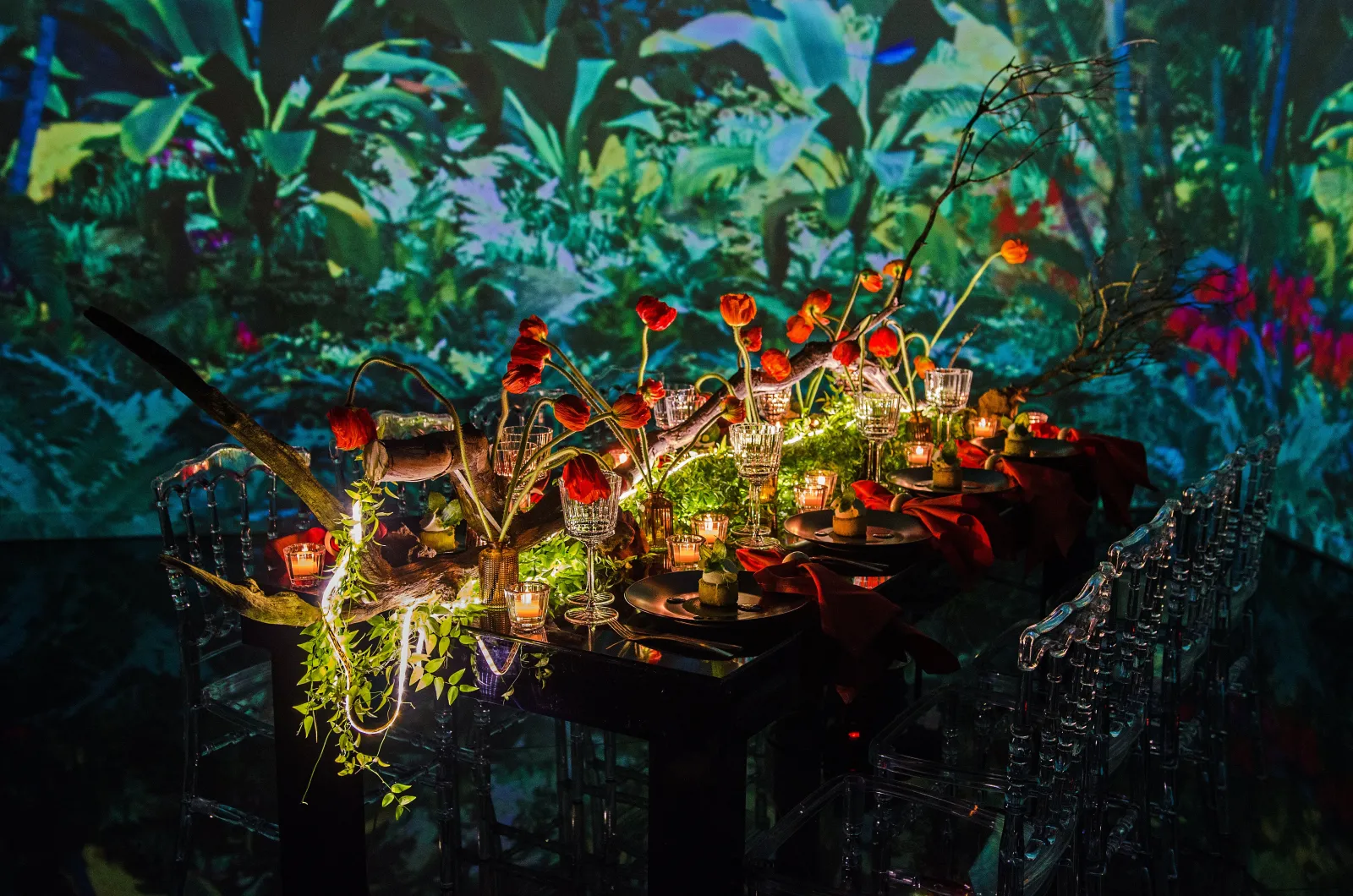 Elegant table setting with red flowers, candles, and greenery under a vibrant botanical projection backdrop