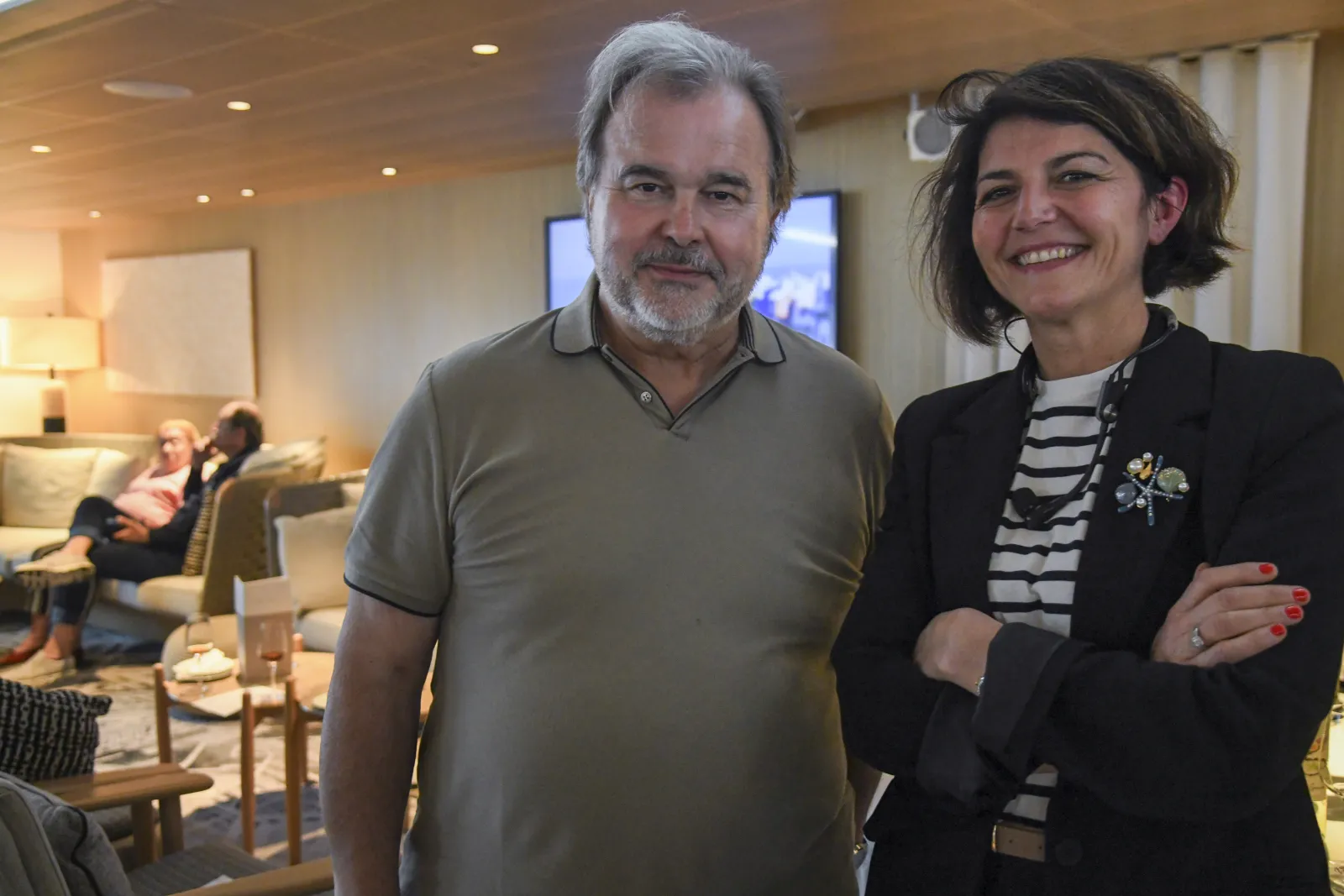 A man in a beige polo shirt and a woman in a black blazer with striped shirt standing together in a modern hotel lounge, smiling at the camera with other guests visible in the background.