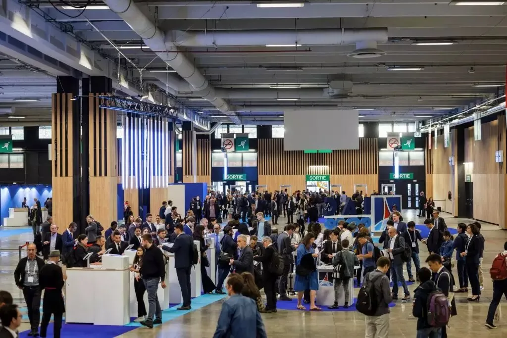 Large indoor convention center or trade show in Bordeaux with numerous attendees in business attire circulating among exhibition booths and information stands under modern ceiling lighting.