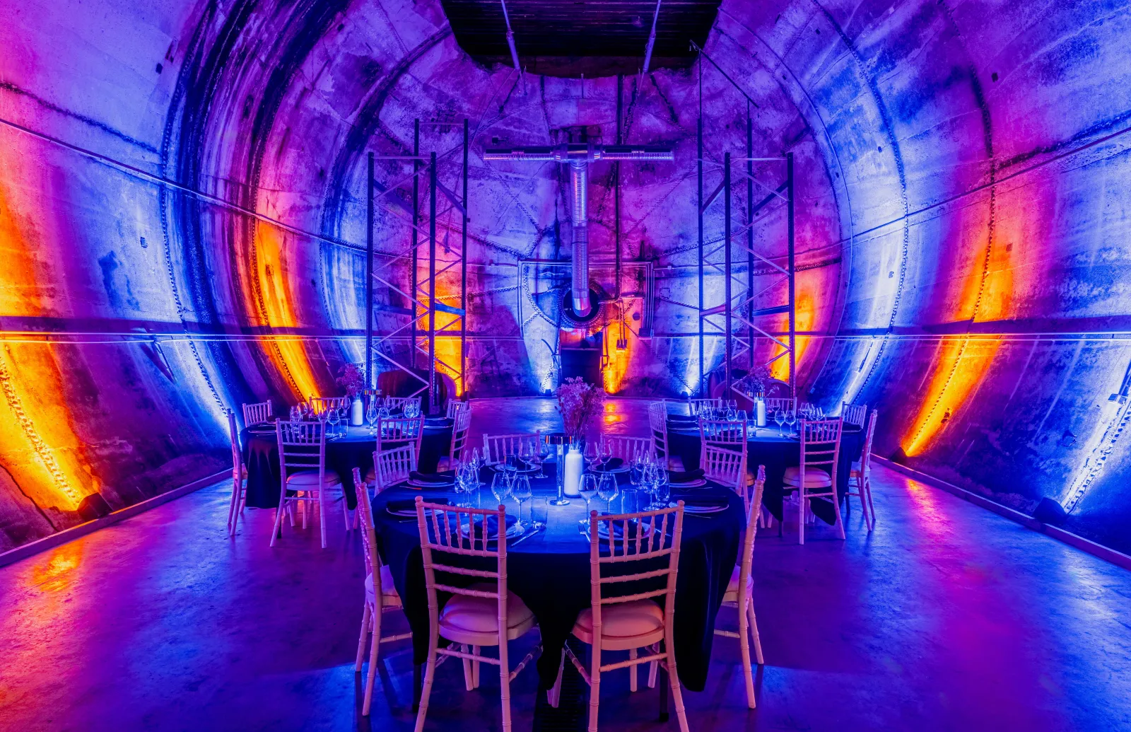 A modern dining space inside an industrial tunnel illuminated with vibrant neon lighting in shades of blue, purple, and orange. Round tables set with place settings and chairs are arranged symmetrically along the tunnel, with metal framework visible overh