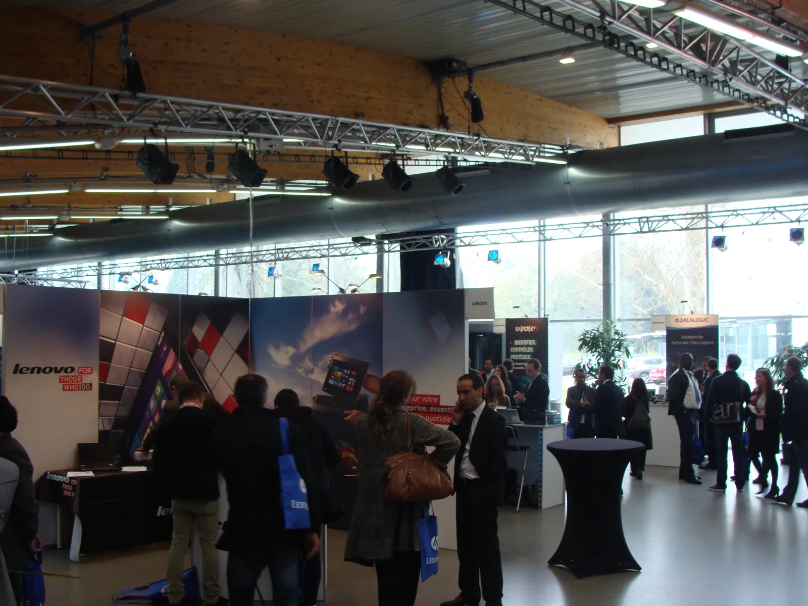 Indoor trade show or exhibition at a modern venue in Bordeaux with attendees in business attire gathered around Lenovo branded booths and displays, featuring large backlit panels and professional lighting equipment suspended from a metal ceiling structure
