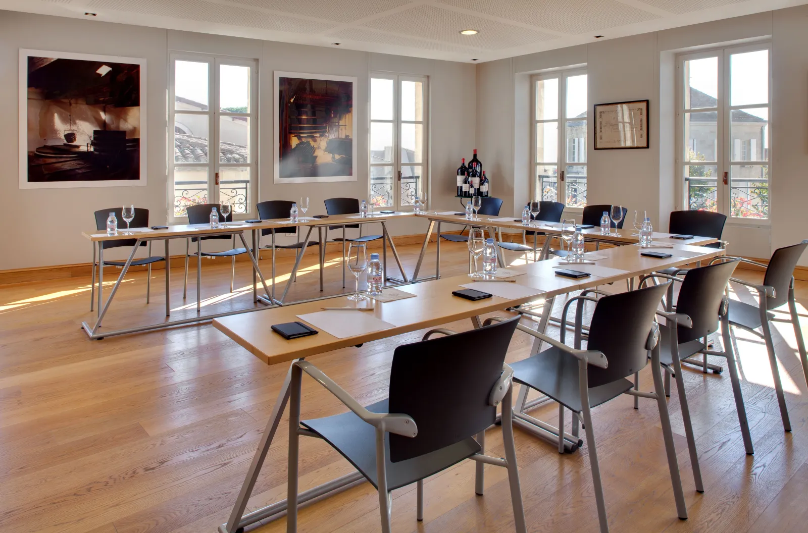 Modern conference room with multiple wooden tables, black chairs, large windows overlooking Bordeaux cityscape, framed artwork on walls, water glasses and notepads on tables, wooden flooring, and contemporary décor.