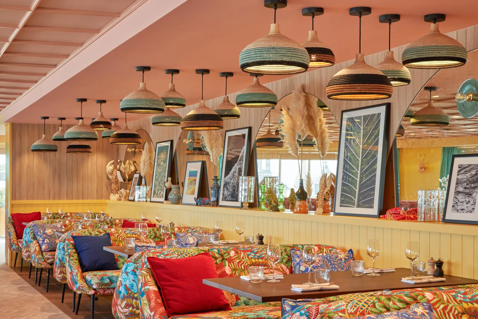 Modern restaurant dining room with coral-pink ceiling, wooden paneling, colorful patterned upholstered chairs, wooden tables, and decorative pendant lights with woven details. Shelving displays artwork and botanical decorations.