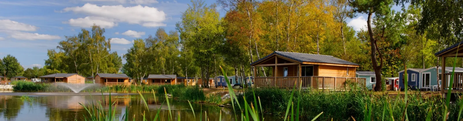 Lakeside camping village with wooden chalets and colorful holiday homes surrounded by birch trees and reeds, reflected in calm water under a partly cloudy sky