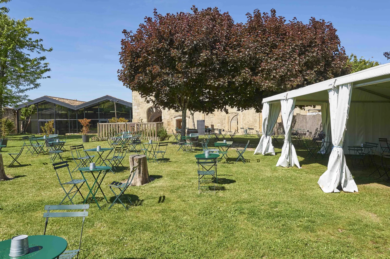 Outdoor courtyard with green lawn, folding chairs, white pergolas with curtains, and historic stone buildings in Bordeaux's Cordeliers district under clear blue sky