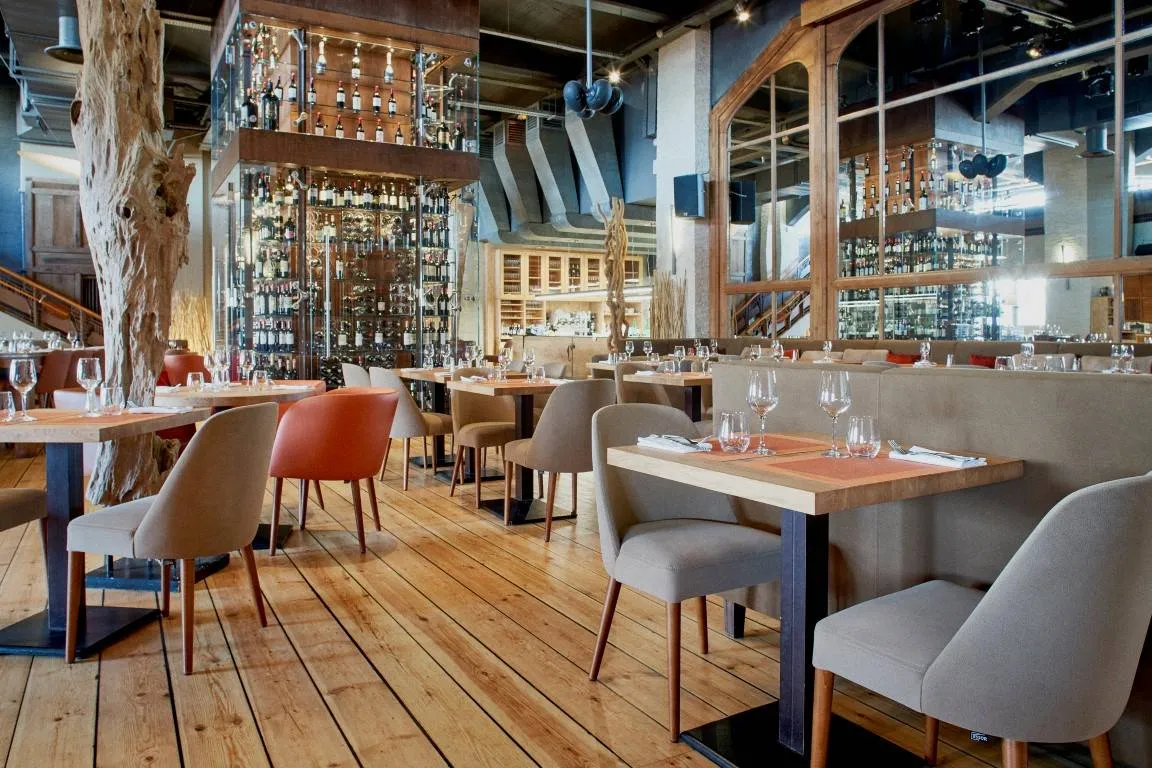 Modern restaurant interior with wooden floors, comfortable beige and red chairs, tables with place settings, extensive bottle display behind glass, and industrial ceiling with exposed beams in Bordeaux Maritime Café.