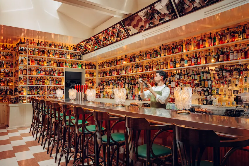 Interior of Osteria Palatino in Bordeaux featuring a long bar counter with a bartender, extensive collection of spirits bottles displayed on illuminated shelves covering all walls, wooden bar stools with red and green seats, and decorative artwork on the 