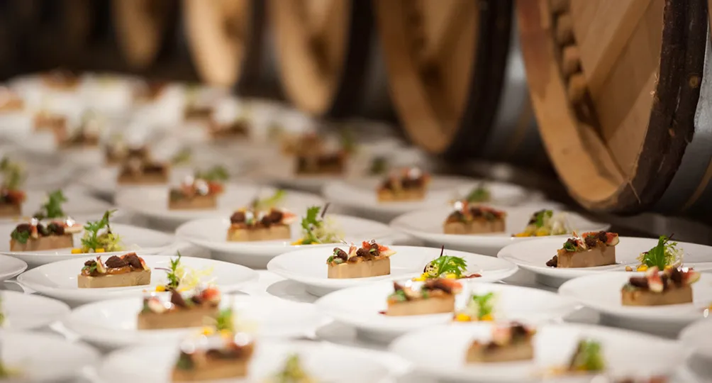 Gourmet canapés on white plates with wooden barrels in the background, representing fine dining cuisine from Bordeaux