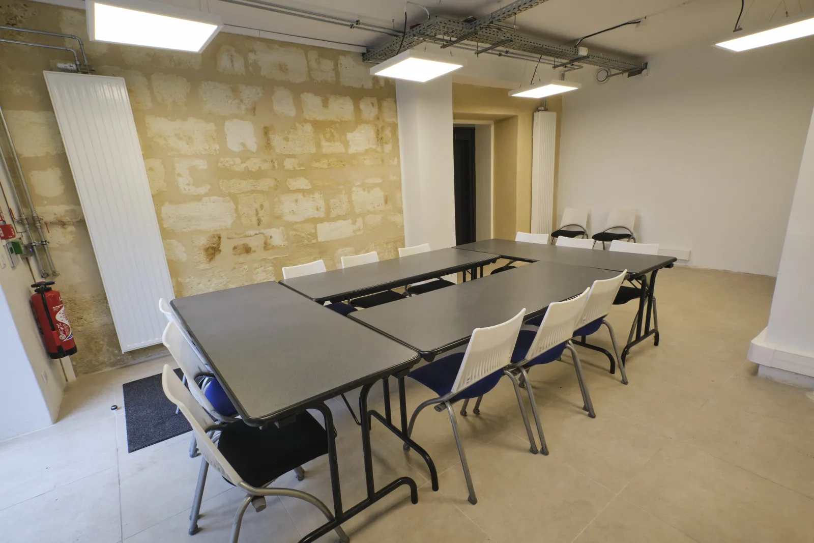 Modern meeting room at ManuCo Bordeaux with modular tables, white chairs, exposed brick wall, LED lighting, and fire safety equipment.